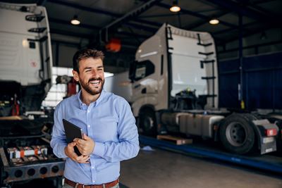 A smiling man in a blue shirt is holding a computer tablet. He is standing in front of transport trucks.